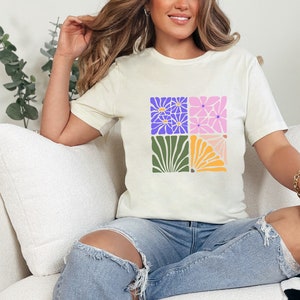 May include: A white t-shirt with a colorful floral design. The design features four squares with different floral patterns in shades of purple, pink, green, and yellow.