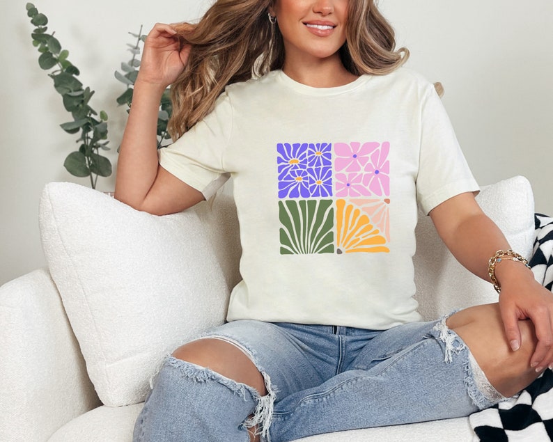 May include: A white t-shirt with a colorful floral design. The design features four squares with different floral patterns in shades of purple, pink, green, and yellow.