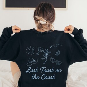May include: Black sweatshirt with a blue graphic design featuring a sun, two wine glasses, a palm tree, sunglasses, a lemon slice, and a hat. The text "Last Toast on the Coast" is printed in blue below the graphic.