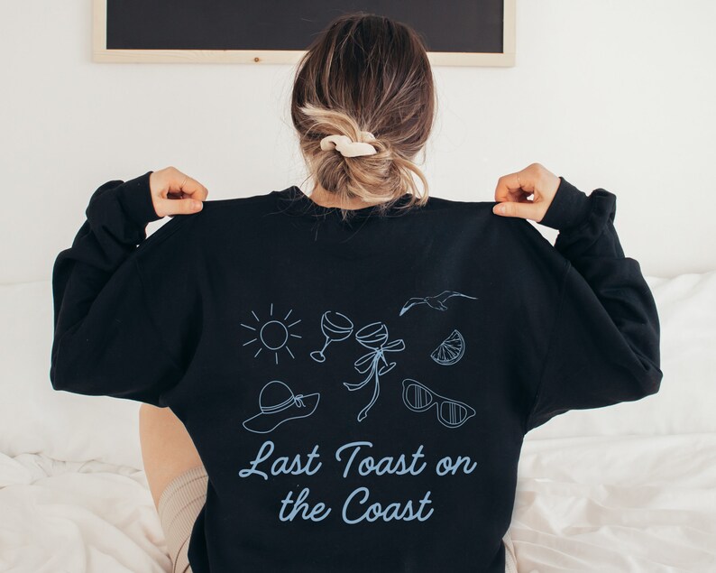 May include: Black sweatshirt with a blue graphic design featuring a sun, two wine glasses, a palm tree, sunglasses, a lemon slice, and a hat. The text "Last Toast on the Coast" is printed in blue below the graphic.