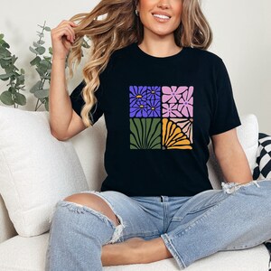 May include: A black t-shirt with a graphic design of four squares featuring different floral patterns in shades of purple, pink, green, and yellow.