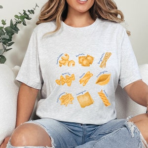May include: A light gray t-shirt with a graphic of different types of pasta, including spaghetti, lasagna, tortellini, and more. The pasta is illustrated in a watercolor style.