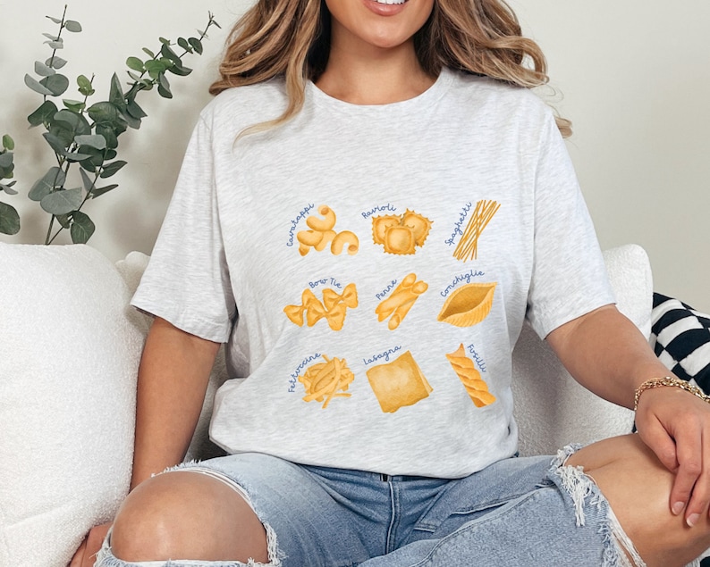 May include: A light gray t-shirt with a graphic of different types of pasta, including spaghetti, lasagna, tortellini, and more. The pasta is illustrated in a watercolor style.