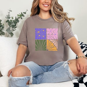 May include: A woman wearing a gray t-shirt with a floral design. The design features four squares with different floral patterns in shades of purple, pink, green, and yellow.