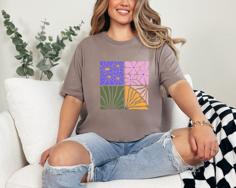 May include: A woman wearing a gray t-shirt with a floral design. The design features four squares with different floral patterns in shades of purple, pink, green, and yellow.