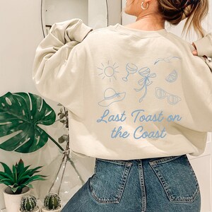 May include: A light beige sweatshirt with a blue graphic design of a sun, two wine glasses, a hat, sunglasses, a lemon, and a seagull. The text "Last Toast on the Coast" is printed in blue below the graphic.