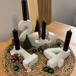 Graue Adventszahlen als Set von 1-4 aus Beton mit schwarzen Kerzen, dekoriert mit Tannenzapfen, Kugeln, Zuckerstangen, kleinen Tannenbäumen auf geflochtenem Untersetzer auf einem Holztisch