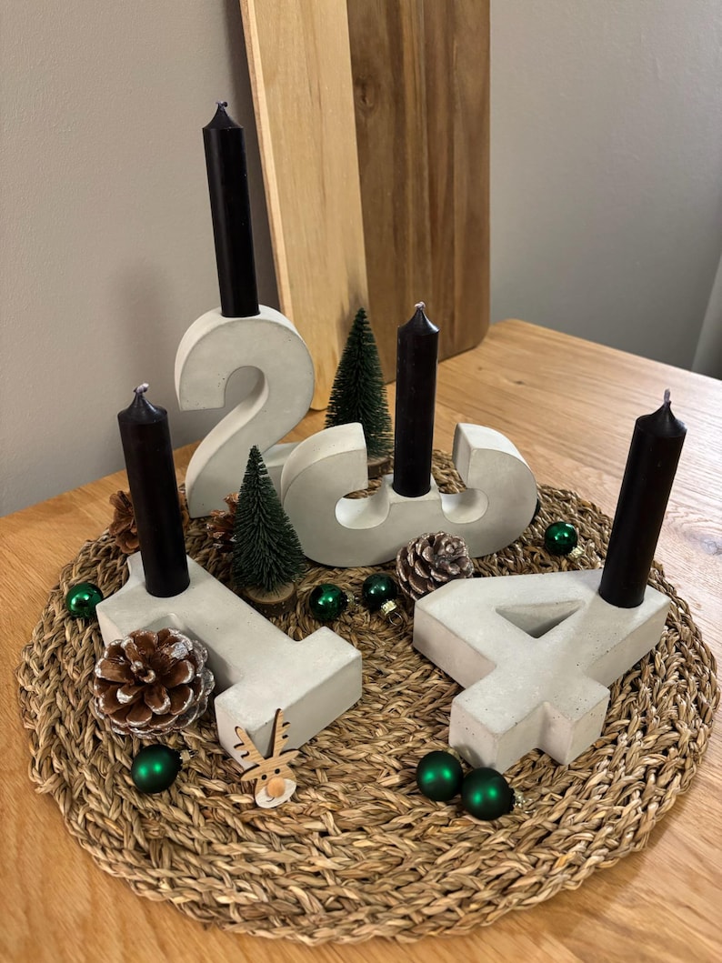 Graue Adventszahlen als Set von 1-4 aus Beton mit schwarzen Kerzen, dekoriert mit Tannenzapfen, Kugeln, Zuckerstangen, kleinen Tannenbäumen auf geflochtenem Untersetzer auf einem Holztisch