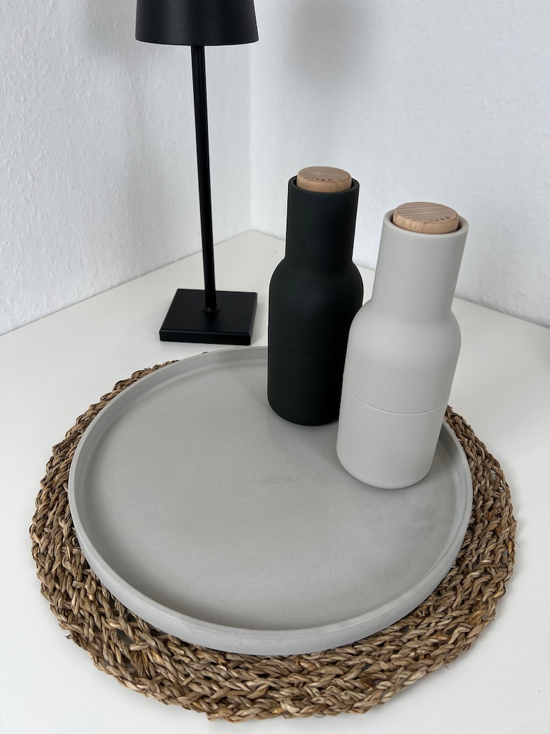 Graues modernes Tablett mit glattem Rand auf einem geflochtenem Untersetzer. Auf dem Tablett steht eine schwarze Pfeffermühle und ein grauer Salzspender. Im Hintergrund steht eine schwarze Stehlampe.