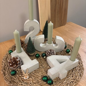 Grauer Adventskranz aus Beton mit Zahlen von 1-4 und Kerzen in Salbei Farbe, dekoriert mit Tannenzapfen und kleinen Weihnachtskugeln, auf geflochtenem Untersetzer auf einem Holztisch