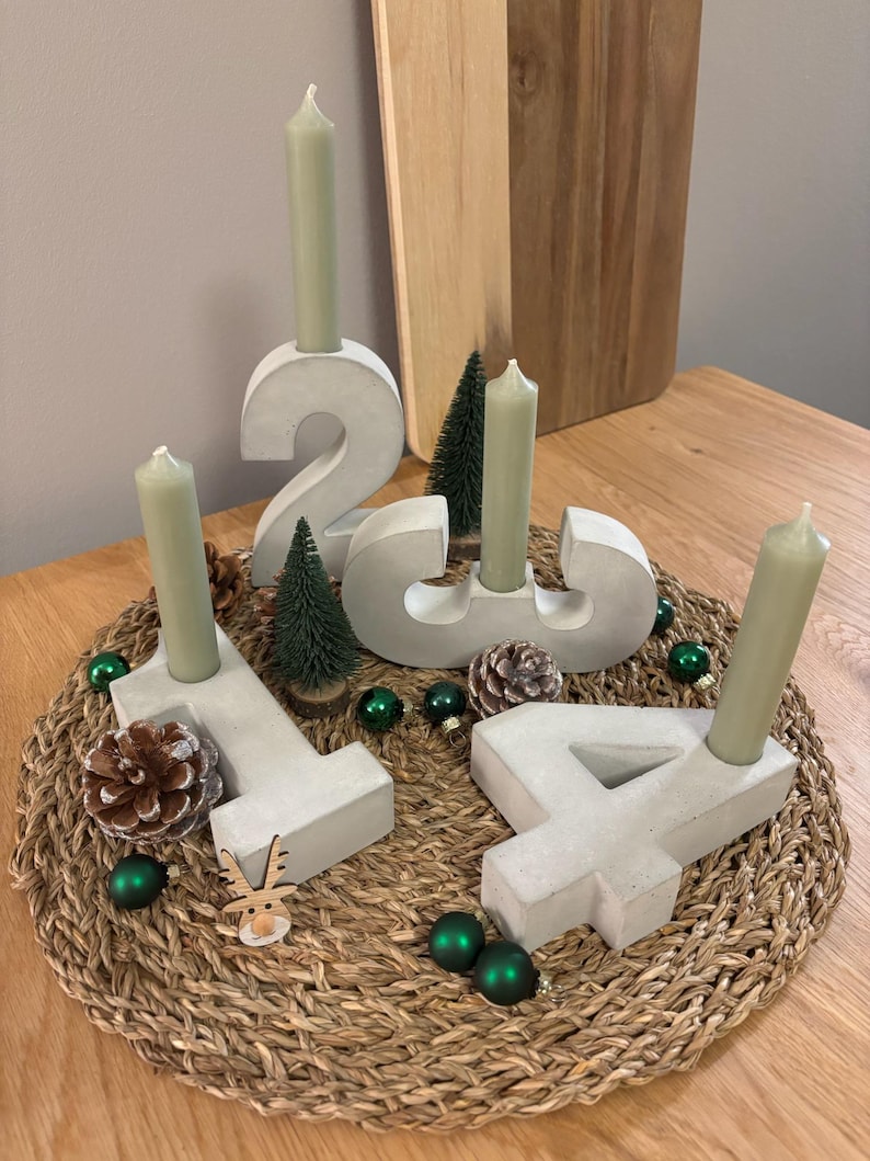Grauer Adventskranz aus Beton mit Zahlen von 1-4 und Kerzen in Salbei Farbe, dekoriert mit Tannenzapfen und kleinen Weihnachtskugeln, auf geflochtenem Untersetzer auf einem Holztisch