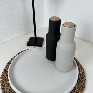 Weißes modernes Tablett mit glattem Rand auf einem geflochtenem Untersetzer. Auf dem Tablett steht eine schwarze Pfeffermühle und ein grauer Salzspender. Im Hintergrund steht eine schwarze Stehlampe.