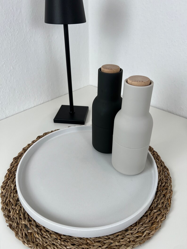Weißes modernes Tablett mit glattem Rand auf einem geflochtenem Untersetzer. Auf dem Tablett steht eine schwarze Pfeffermühle und ein grauer Salzspender. Im Hintergrund steht eine schwarze Stehlampe.