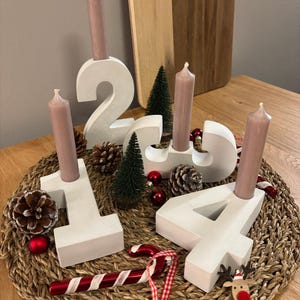 Adventskranz aus Beton mit weißen Zahlen von 1-4 mit taupefarbenen Kerzen, dekoriert mit Tannenzapfen, Kugeln, Zuckerstangen, auf geflochtenem Untersetzer auf einem Holztisch