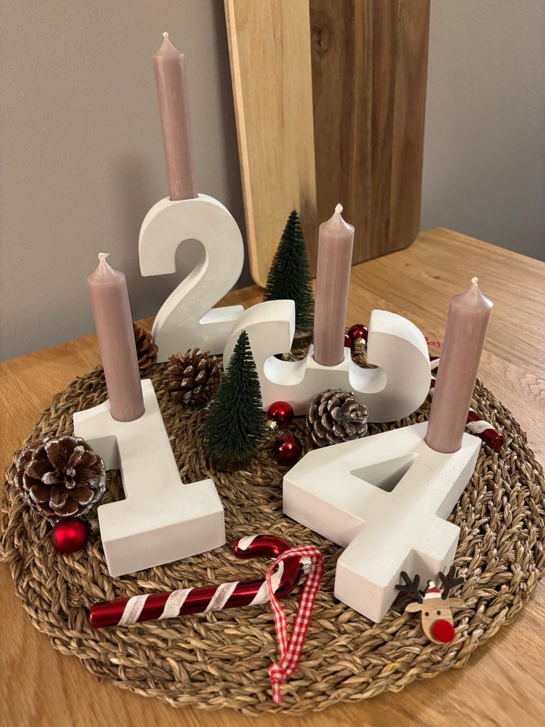 Adventskranz aus Beton mit weißen Zahlen von 1-4 mit taupefarbenen Kerzen, dekoriert mit Tannenzapfen, Kugeln, Zuckerstangen, auf geflochtenem Untersetzer auf einem Holztisch
