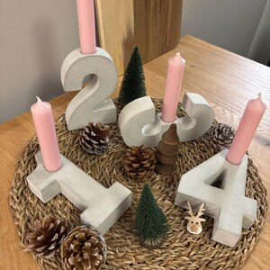 Graue Adventszahlen als Set von 1-4 aus Beton mit rosafarbenen Kerzen, dekoriert mit Tannenzapfen, und kleinen Tannenbäumen auf geflochtenem Untersetzer auf einem Holztisch