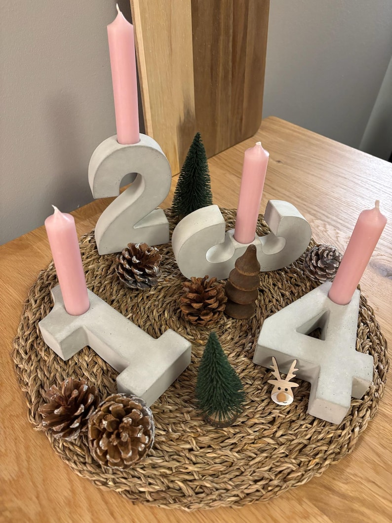 Graue Adventszahlen als Set von 1-4 aus Beton mit rosafarbenen Kerzen, dekoriert mit Tannenzapfen, und kleinen Tannenbäumen auf geflochtenem Untersetzer auf einem Holztisch