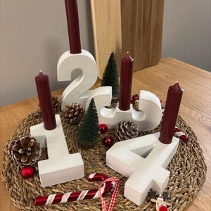 Weißer Adventskranz aus Beton mit dunkelroten Kerzen, dekoriert mit Tannenzapfen, Kugeln, Zuckerstangen, auf geflochtenem Untersetzer mit einem Holzbrettchen im Hintergrund auf einem Holztisch