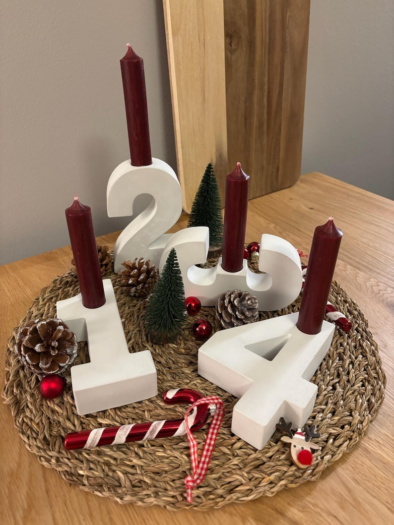 Weißer Adventskranz aus Beton mit dunkelroten Kerzen, dekoriert mit Tannenzapfen, Kugeln, Zuckerstangen, auf geflochtenem Untersetzer mit einem Holzbrettchen im Hintergrund auf einem Holztisch