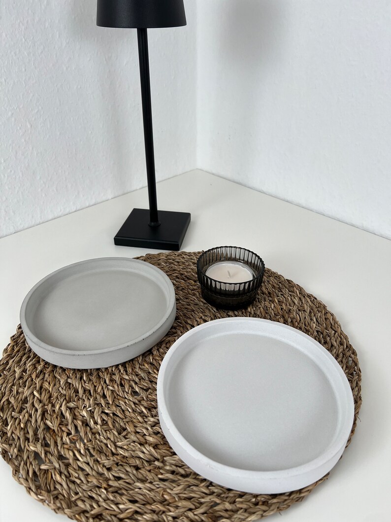 Ein graues und ein weißes kleines Tablett nebeneinander auf einem geflochtenem Untersetzer. Daneben ein schwarzer Teelichthalter aus Glas mit einem Teelicht. Im Hintergrund steht eine schwarze Lampe