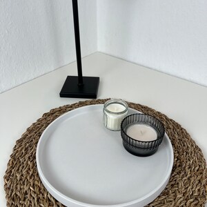 Weißes modernes Tablett mit glattem Rand auf einem geflochtenem Untersetzer. Auf dem Tablett stehen zwei Kerzen in einem schwarzen und durchsichtigen Glas. Im Hintergrund steht eine schwarze Stehlampe.