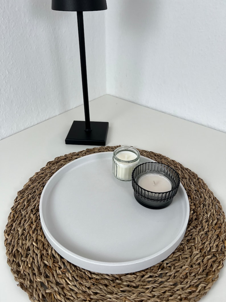 Weißes modernes Tablett mit glattem Rand auf einem geflochtenem Untersetzer. Auf dem Tablett stehen zwei Kerzen in einem schwarzen und durchsichtigen Glas. Im Hintergrund steht eine schwarze Stehlampe.