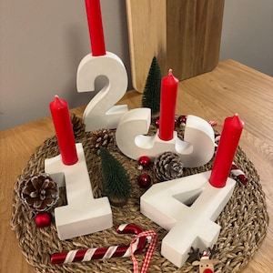 Adventskranz aus Beton mit weißen Zahlen von 1-4 mit roten Kerzen, dekoriert mit Tannenzapfen, Kugeln, Zuckerstangen, auf geflochtenem Untersetzer auf einem Holztisch