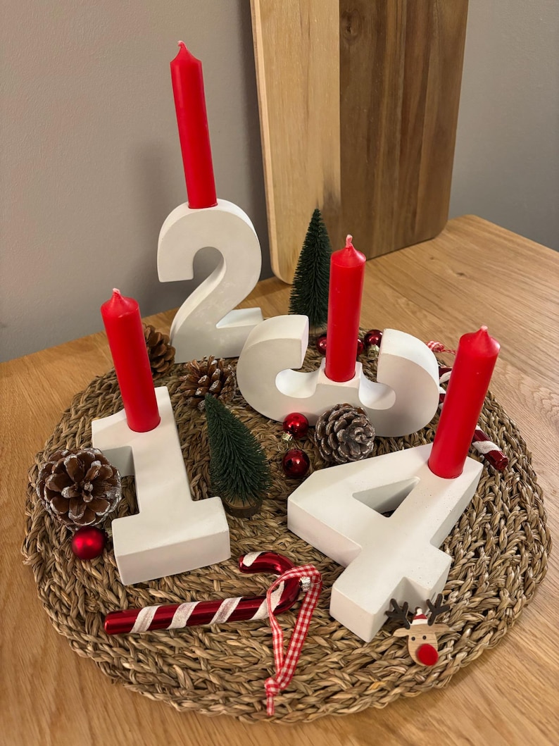 Adventskranz aus Beton mit weißen Zahlen von 1-4 mit roten Kerzen, dekoriert mit Tannenzapfen, Kugeln, Zuckerstangen, auf geflochtenem Untersetzer auf einem Holztisch