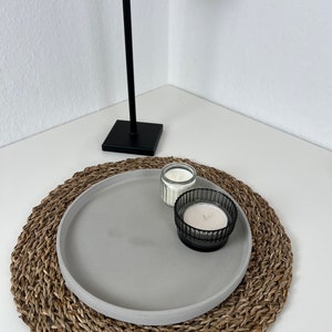 Graues modernes Tablett mit glattem Rand auf einem geflochtenem Untersetzer. Auf dem Tablett stehen zwei Kerzen in einem schwarzen und durchsichtigen Glas. Im Hintergrund steht eine schwarze Stehlampe.