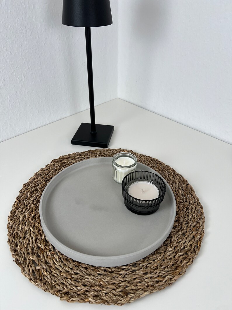 Graues modernes Tablett mit glattem Rand auf einem geflochtenem Untersetzer. Auf dem Tablett stehen zwei Kerzen in einem schwarzen und durchsichtigen Glas. Im Hintergrund steht eine schwarze Stehlampe.