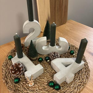 Moderne graue Zahlen aus Beton dekoriert als Adventskranz auf einem geflochtenem Untersetzer. Der Adventskranz steht auf einem Holztisch mit Deko aus Tannenzapfen, kleinen Tannenbäumen und Weihnachtskugeln.