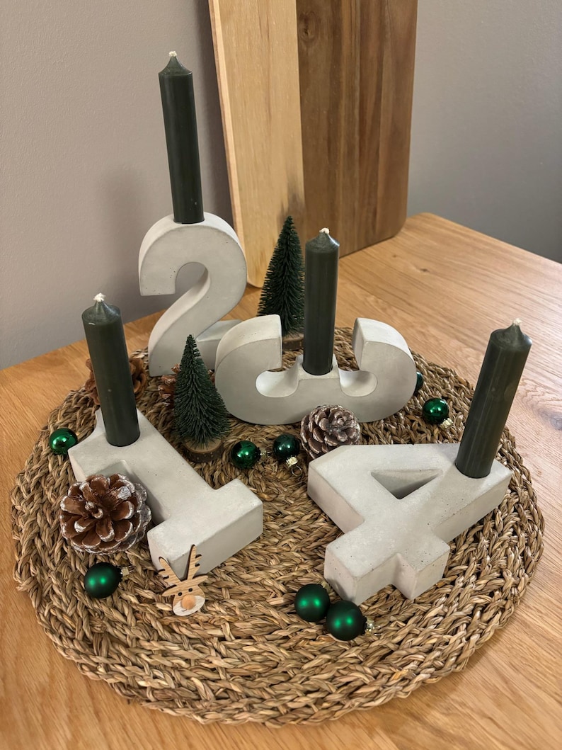 Moderne graue Zahlen aus Beton dekoriert als Adventskranz auf einem geflochtenem Untersetzer. Der Adventskranz steht auf einem Holztisch mit Deko aus Tannenzapfen, kleinen Tannenbäumen und Weihnachtskugeln.