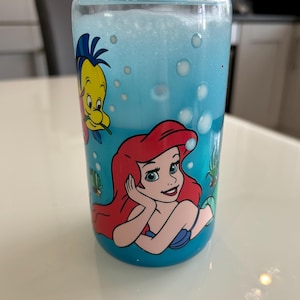May include: A clear glass tumbler with a screw-on lid, filled with blue liquid and bubbles. The tumbler features a colourful graphic of Ariel and Flounder from The Little Mermaid. The design is set against a blue background.