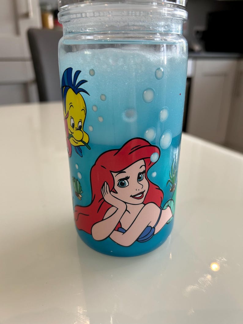May include: A clear glass tumbler with a screw-on lid, filled with blue liquid and bubbles. The tumbler features a colourful graphic of Ariel and Flounder from The Little Mermaid. The design is set against a blue background.