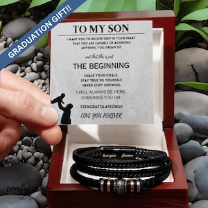 Puede incluir: Una caja de madera con un brazalete de cuero negro en su interior. La caja tiene una tarjeta dentro que dice "A mi hijo" e incluye un mensaje sobre creer en ti mismo y perseguir tus metas. La tarjeta también dice "¡Felicidades! Te quiero para siempre".