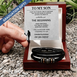 Puede incluir: Un brazalete de cuero negro con cierre plateado y detalles dorados se presenta en una caja de madera con una tarjeta que dice "A mi hijo" e incluye un mensaje de aliento y felicitaciones.