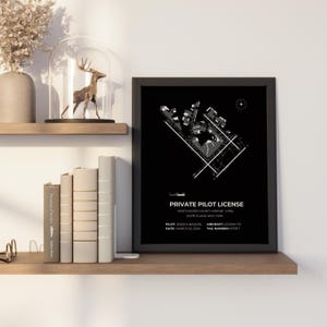 May include: A framed black and white print labeled "Private Pilot License" with a diagram of an airport. The text includes the pilot's name, aircraft, and date. The print is displayed on a wooden shelf with books and decorative items.