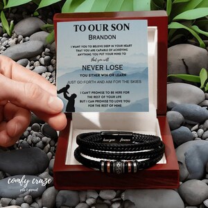 Puede incluir: Una caja de madera con un brazalete de cuero negro en su interior. La caja tiene una tarjeta con un mensaje que dice: "A nuestro hijo Brandon, quiero que creas en lo profundo de tu corazón que eres capaz de lograr cualquier cosa que te propongas. Nunca pierdas. O ganas o aprendes. Solo avanza y apunta a las estrellas. No puedo prometerte estar aquí por el resto de tu vida, pero puedo prometerte amarte por el resto de la mía."