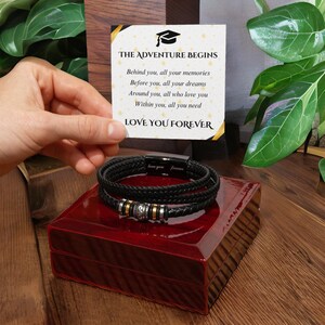 Puede incluir: Un brazalete de cuero negro con un dije plateado y dorado. El brazalete está en una caja roja con una tarjeta que dice "The Adventure Begins" y "Love You Forever".