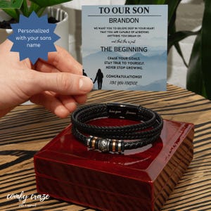 Puede incluir: Una pulsera de cuero negro con detalles plateados y dorados descansa sobre una caja de madera roja. Una tarjeta con el texto "TO OUR SON BRANDON" y un mensaje se sostiene sobre la pulsera. La pulsera está grabada con "love you forever".