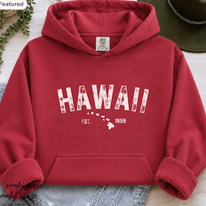 May include: A red hooded sweatshirt featuring a white distressed graphic that reads HAWAII with a small map of the Hawaiian islands and the text EST. 1959. The hoodie has a front kangaroo pocket, long sleeves with ribbed cuffs, and a Comfort Colors label.