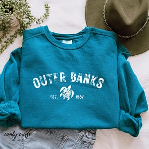 Puede incluir: Sudadera azul turquesa con un gráfico blanco de una tortuga marina y el texto "OUTER BANKS EST. 1567".