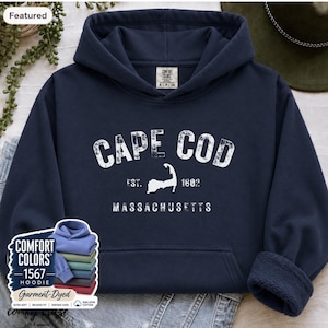 May include: A navy blue hooded sweatshirt featuring white distressed text that reads CAPE COD EST. 1602 MASSACHUSETTS with a graphic of the Cape Cod peninsula. The hoodie has a front kangaroo pocket, long sleeves with ribbed cuffs, and a Comfort Colors label.