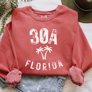 May include: A red-orange sweatshirt with the text "30A FLORIDA" in white, along with a palm tree graphic. The sweatshirt is folded on top of a pair of blue denim shorts. The sleeves are cuffed.