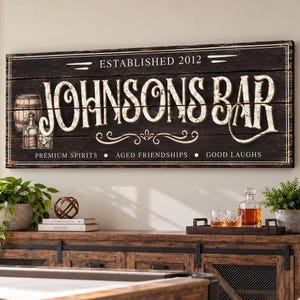 May include: A rectangular wooden wall sign with a dark brown finish and distressed white lettering. The text reads "ESTABLISHED 2012", "JOHNSONS BAR", and "PREMIUM SPIRITS • AGED FRIENDSHIPS • GOOD LAUGHS". It features a graphic of a barrel and glass.