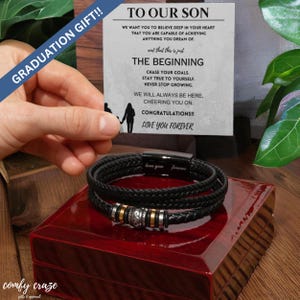 Puede incluir: Un brazalete de cuero negro con cierre plateado y cuentas plateadas. El brazalete está en una caja roja con una tarjeta que dice "A nuestro hijo" e incluye un mensaje de felicitación y amor.