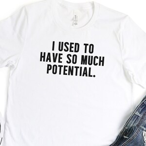 May include: White t-shirt with the text "I USED TO HAVE SO MUCH POTENTIAL" in black. The shirt is paired with blue denim jeans and black canvas sneakers with white soles and laces. The sneakers are placed near the shirt and jeans.
