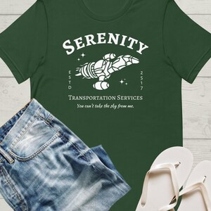 May include: A green t-shirt with a white graphic of a spaceship with the text "Serenity" above it and "Transportation Services" below it. The text "ESTD 2517" is on the left side of the spaceship and the text "You can't take the sky from me." is below the text "Transportation Services".