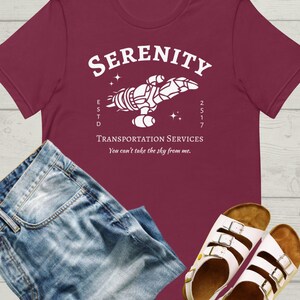 May include: A maroon t-shirt with a white graphic of a spaceship with the text "Serenity" above it and "ESTD 2517" below it. The text "Transportation Services" is below the spaceship and the text "You can't take the sky from me." is below that.