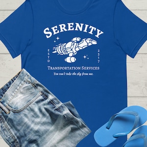 May include: A royal blue t-shirt with a white graphic of a spaceship with the text "Serenity" above it and "Transportation Services" below it. The text "Est'd 2517" is on the left side of the spaceship and the text "You can't take the sky from me." is below the text "Transportation Services".